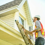 A man with a white hard hat holding a clipboard, inspect house